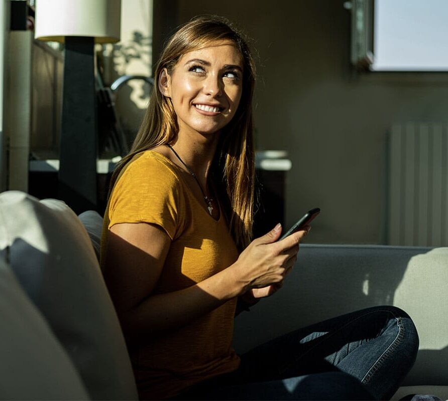 A woman sitting on the couch with her phone in her hands smiling up beside her.