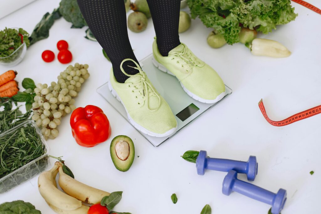 Someone standing on a scale in yellow-green runners with two dumbbells and lots of different vegetables lying around the scale on the ground.