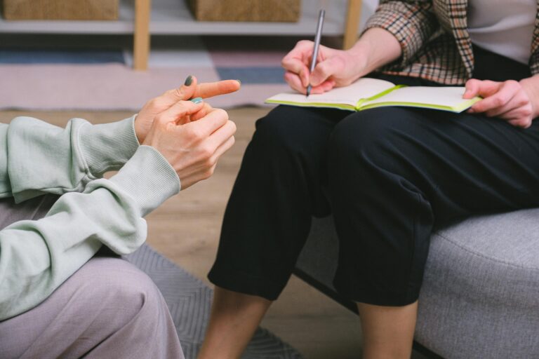 A medical consultation between a patient and a healthcare provider. The shot shows the two people facing each other chest down, with the healthcare provider making notes in a notebook.