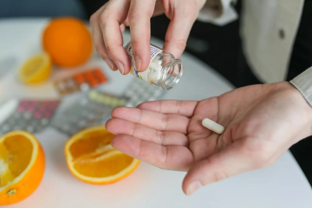 Someone taking a supplement out of a jar with oranges and other supplements behind them on the table.